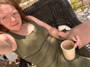 Sitting in a green dress awkwardly in a chair. Red hair, coffee cup in hand and one tattoo visible of a collection of semicolons in pride colors. 