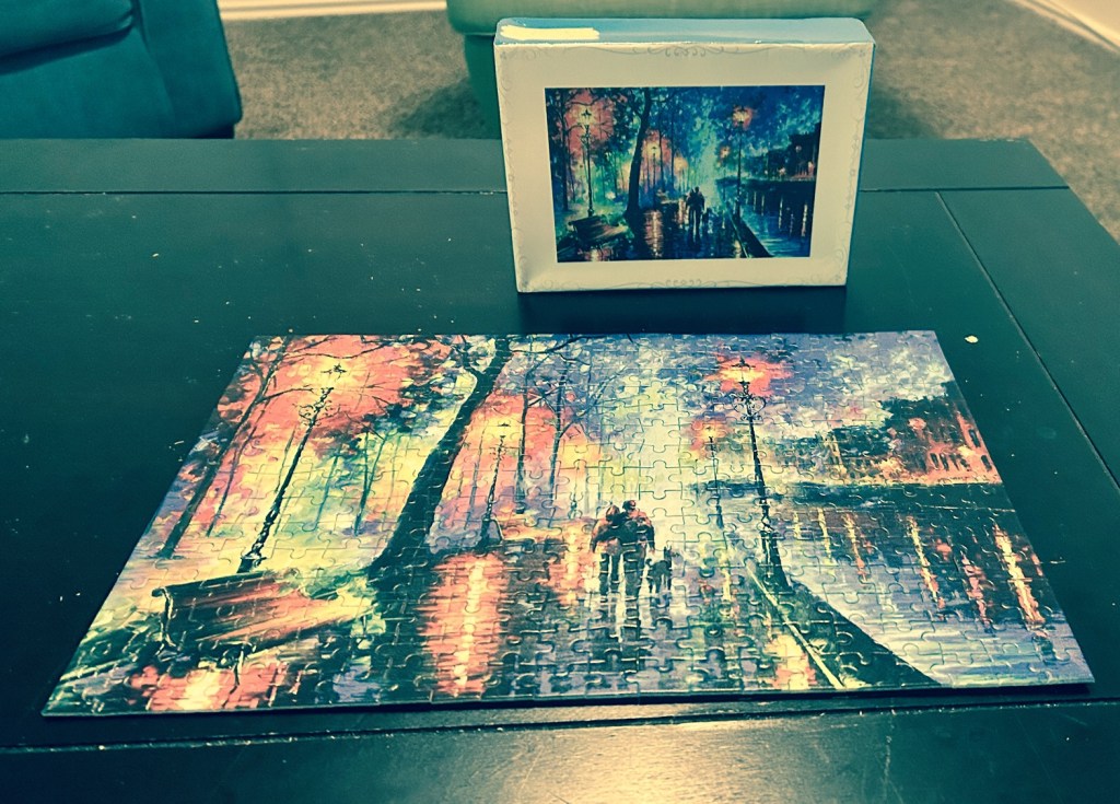 A completed jigsaw puzzle depicting a colorful street scene with lampposts and people walking, displayed on a table, accompanied by its puzzle box showing the same image.