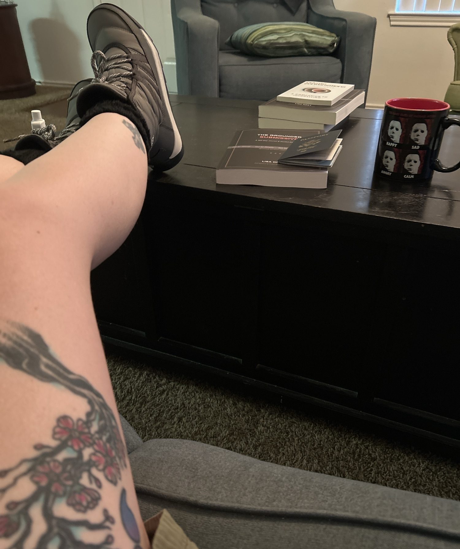 First-person view of legs resting on a coffee table wearing grey waterproof boots. A passport rests nearby next to a coffee mug featuring Michael Myers' mask with happy, sad, angry, and calm expressions.